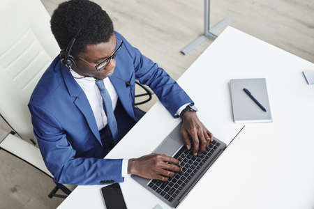 High angle view of African businessman in headphones typing on laptop and communicating online at...