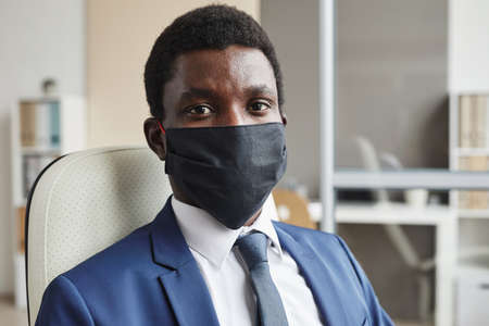 Portrait of African young businessman in protective mask looking at camera while sitting at office