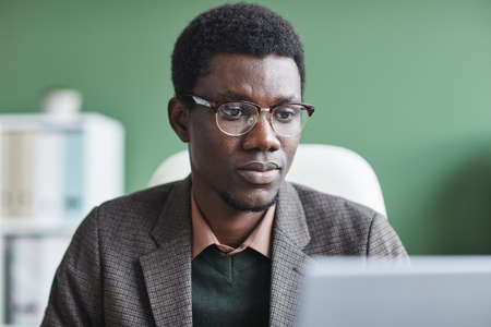 African manager in eyeglasses looking at monitor of laptop during his work at office