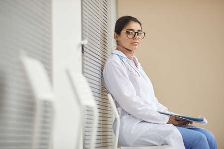 Portrait of young nurse in white coat looking at camera while sitting at the corridor of hospitalの写真素材
