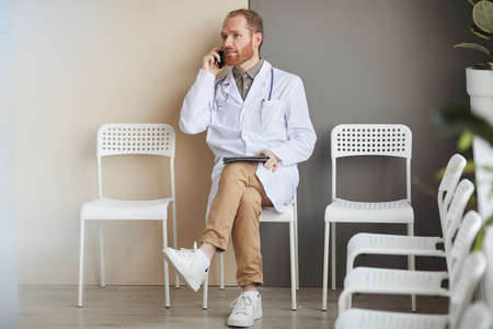Male doctor in white coat sitting on chair talking on mobile phone and using tablet pc during his...