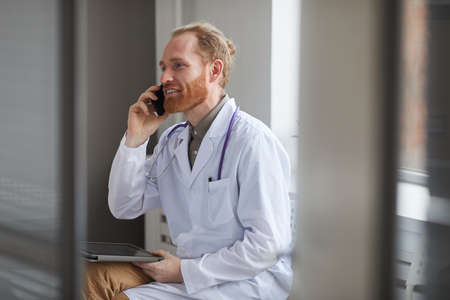 Confident doctor in white coat has a conversation on mobile phone while sitting at the corridorの写真素材