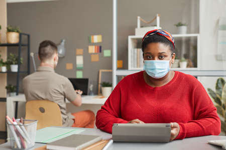 Portrait of African overweight woman in protective mask looking at camera while sitting at workplace...