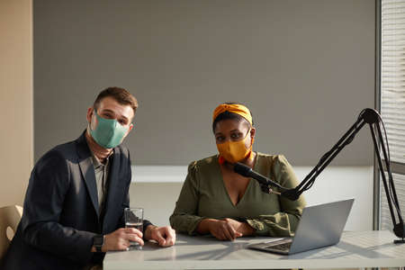 Portrait of African radio dj and guest sitting at the table in protective mask and looking at camera during interview on radioの写真素材