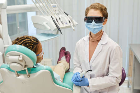 Portrait of young dentist in protective glasses and mask treating patients teeth at dental clinic