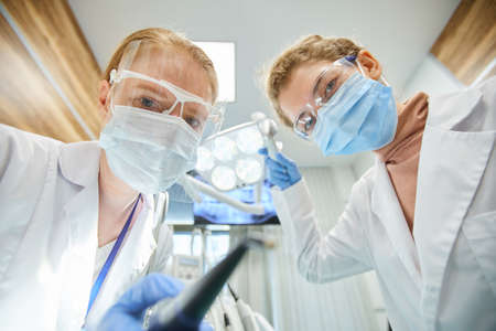 Portrait of two dentists in protective masks and glasses looking at camera while working with...