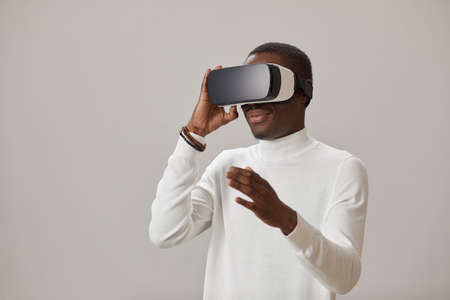 African young man wearing VR glasses and playing in virtual game against the gey background