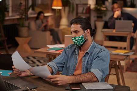 Young businessman in casual clothing working in protective mask at his table with documents at officeの写真素材