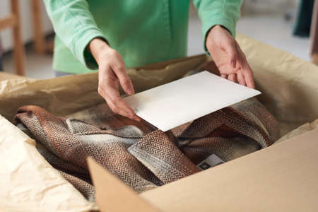 Close-up of woman packing new clothes in box and putting the postcard on it before delivering