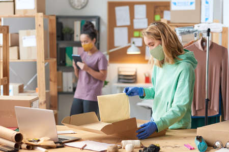 Young woman in mask packing parcel while standing near the table with her colleague in the background in workshopの写真素材