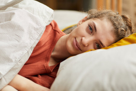 Portrait of young woman lying on pillow looking at camera while resting in her bed at home