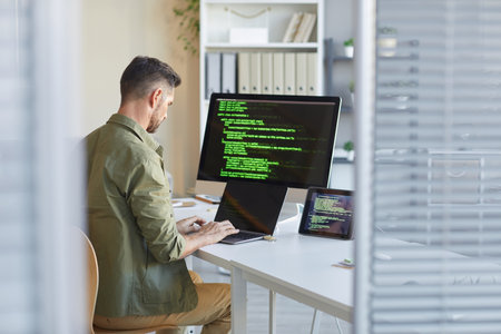 Mature technician sitting at his workplace in front of computer monitor and typing on laptop at IT officeの写真素材