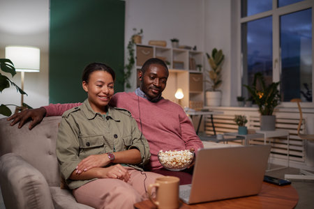 Young couple sitting on sofa with popcorn and watching an interesting movie on laptop at homeの写真素材
