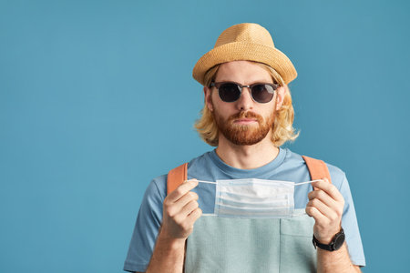 Portrait of tourist in sunglasses and in hat wearing protective mask isolated on blue background