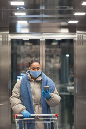 Young woman in protective mask standing in elevator with shopping cart and making selfie on the...