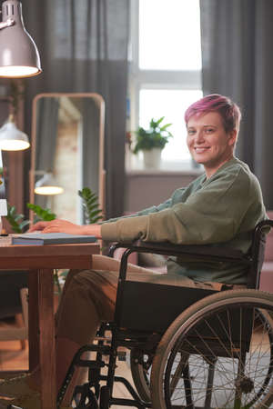 Portrait of young woman sitting in wheelchair and looking at camera while working on computer at...
