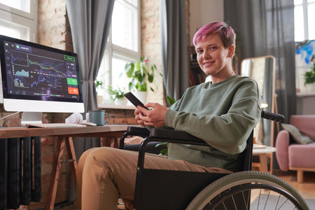 Disabled woman with short hair sitting on wheelchair and working online on mobile phone during her work on computer at homeの写真素材