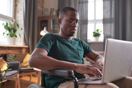 African disabled man sitting in wheelchair with laptop on his knees and typing on it working online...