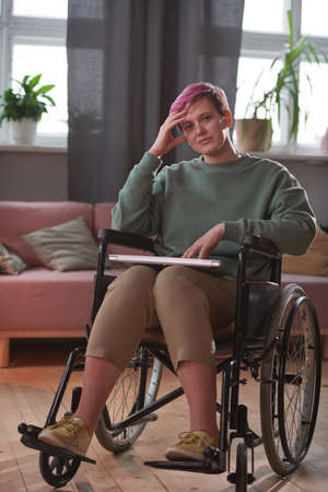 Young woman with short hair sitting in wheelchair with laptop looking at camera in the room at home