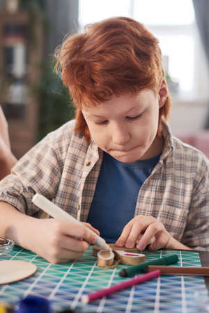 Boy sitting at the table and decorating craft with crayons during art lesson