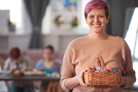 Portrait of young woman holding basket with Easter eggs and smiling at camera with children in the...