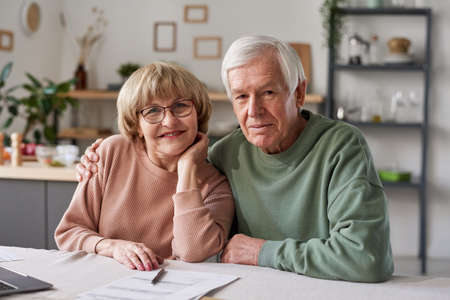 Portrait of senior couple embracing and smiling at camera while sitting at the table with laptop and documents at homeの写真素材