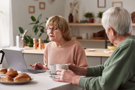 Senior woman concentrating on her online work on laptop while having breakfast together with her...