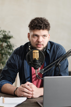 Young male radio dj sitting at the table with laptop and speaking in microphone during his work