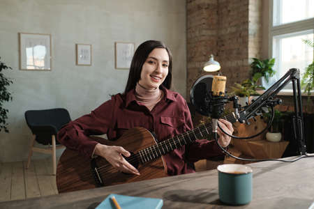 Portrait of young woman smiling at camera while playing guitar during air on the radioの写真素材