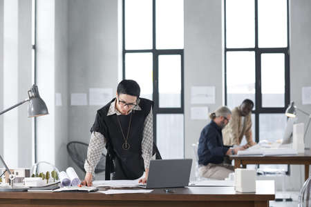Architect working at the table with laptop and blueprints with her colleagues in the background