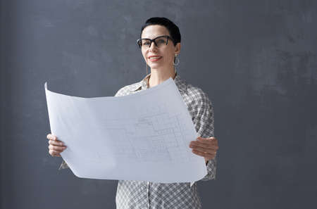Portrait of mature architect in eyeglasses examining blueprint and looking at camera against the...
