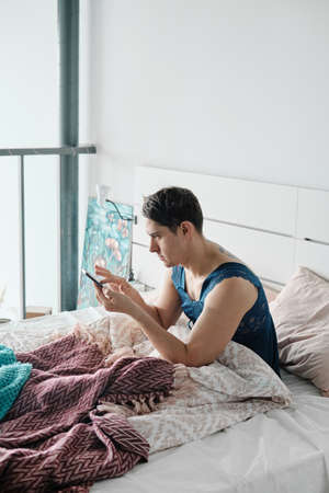 Transgender in female pyjamas texting a message on his mobile phone while sitting in the bed in the morningの写真素材