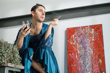 Transgender in female clothes sitting with glass of wine and smoking a cigarette at home