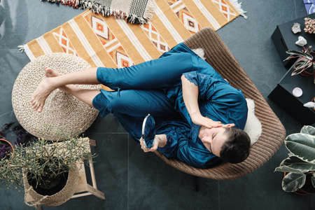High angle view of young man in female dressing gown sitting on armchair and looking at mirror he caring about his beautyの写真素材