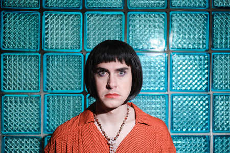 Portrait of transsexual in short black wig looking at camera standing against the blue glass wall