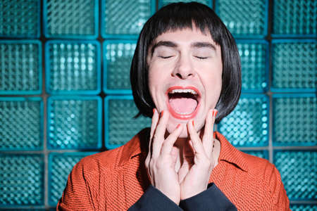 Young transgender in short black wig opening his mouth with red lipstick posing at camera against...