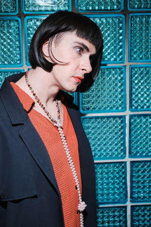 Young transgener in short black wig dress and necklace posing against the blue glass wall
