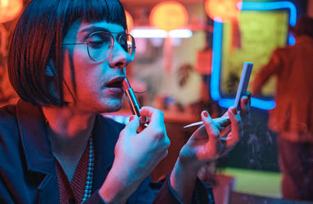 Young man in short black wig applying lipstick and looking at mirror while sitting in the nightclub