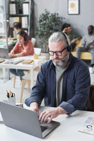 Mature man in headphones working on laptop at his workplace during his work in call center