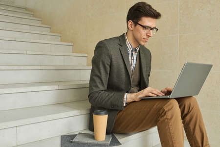 Young businesswoman in eyeglasses sitting on stairs and typing on laptop computer he working online