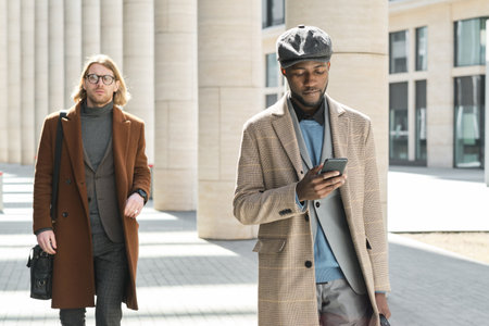 African young man using mobile phone while walking along the street with man in the background