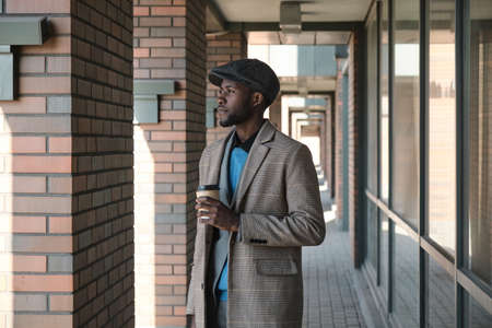 African young businessman holding cup of coffee and looking away standing on the streetの写真素材