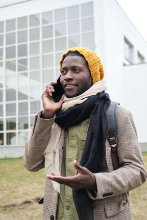 African young man in hat talking on mobile phone while standing in the cityの写真素材