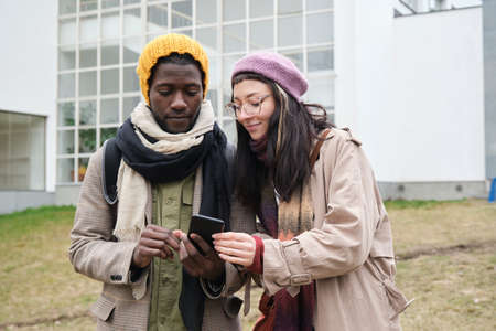 Young couple in warm clothing using mobile phone together outdoorsの写真素材