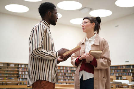 Two students discussing books and studying while standing in the libraryの写真素材