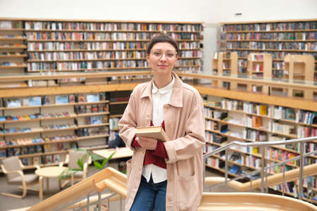 Portrait of young woman with book standing in the big libraryの写真素材