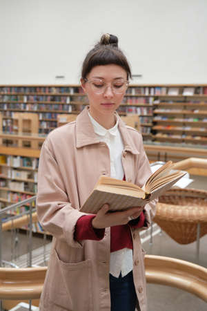 Young woman in eyeglasses reading a book while standing in the big library