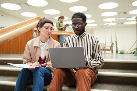 African man using laptop and studying together with woman while they sitting on stairs in the...