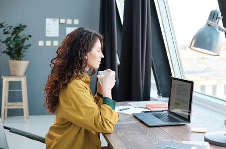 Young woman drinking coffee while sitting at the table in front of laptop and working at home