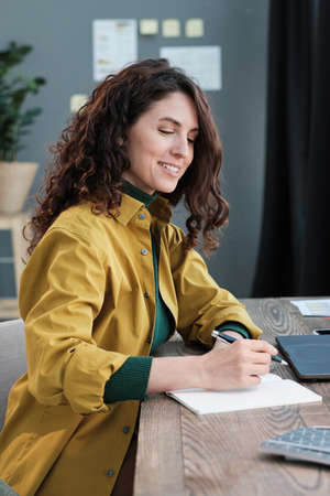 Young businesswoman sitting at the table and writing in documents at the table